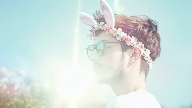 A young man stands outdoors during spring, showcasing a light-hearted look with Easter bunny ears and a floral crown. The bright sky and greenery add to the cheerful atmosphere