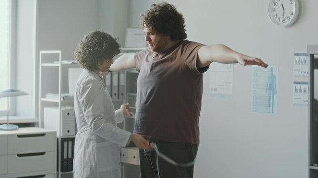 Female doctor measuring waist of obese man with tape measure and speaking with him during medical checkup in clinic