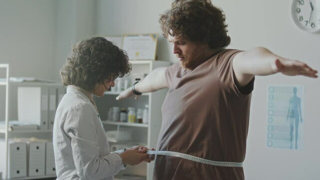 Female doctor measuring body circumference of obese man, then giving supportive words and advice on weight loss during health checkup in medical office