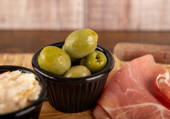 A wooden platter of food with a black bowl of olives and ham. The olives are green and the ham is pink