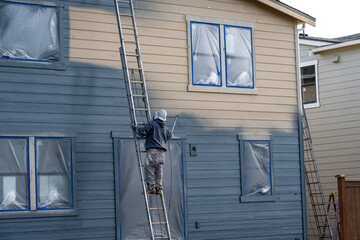 Professional painter in hard hat and air respirator on extension ladder with paint sprayer painting...