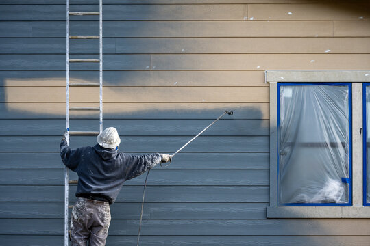 Professional painter in hard hat and air respirator on extension ladder with paint sprayer painting back exterior of house, job site for new housing development
