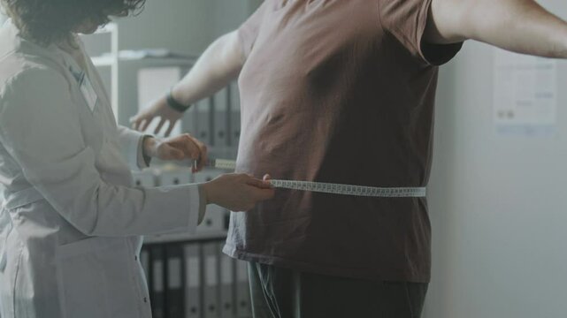Female dietitian in white coat measuring waist circumference of obese male patient with tape measure during medical checkup in clinic