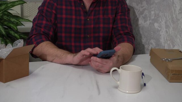 A man uses a phone while sitting at a table with parcels. The guy taps on the smartphone screen when placing an order or making a refund in the app.