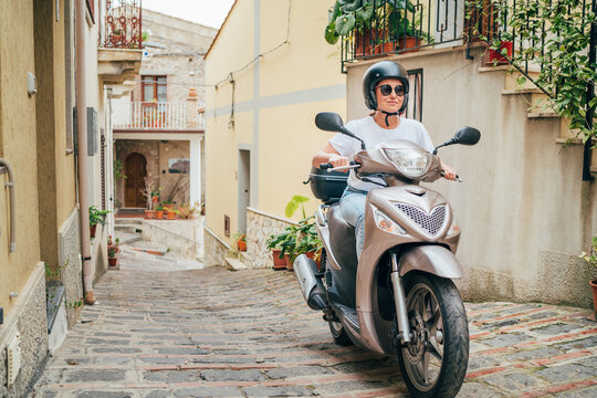 Fototapeta Cheerfully smiling woman in helmet and sunglasses fast riding the moto scooter on the narrow Sicilian old town streets. Happy Italian vacation and transportation concept.