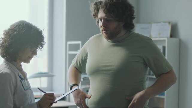 Overweight male patient touching his waist and talking to female doctor writing down notes on clipboard during medical consultation in clinic. Tilt-up shot