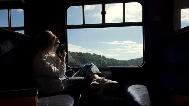 girl sits in a retro train and films the view from the window on her phone. A young woman records the landscape outside the window of a vintage passenger carriage on her smartphone camera.