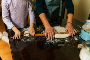 Grandmother teaching her granddaughter how to make dough using rolling pins on a kitchen table,...