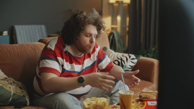 Young man with obesity sitting on couch in cozy living room, eating fast food such as chips and French fries with sauce while watching TV at home