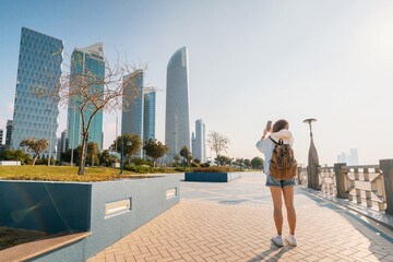 Young woman taking photos of modern skyscrapers with smartphone in Abu Dhabi, United Arab Emirates
