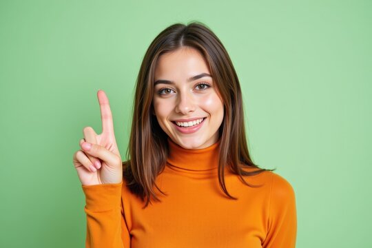A Smiling Young Caucasian Woman in an Orange Turtleneck Sweater Holding Up Her Index Finger Against a Vibrant Green Background, Signifying Positivity