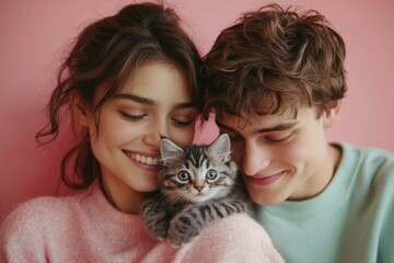 A young couple cuddling a cute tabby kitten against a pink background.
