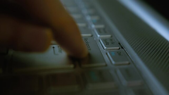 Close-up of a person's finger pressing the backspace key on a laptop keyboard in a dimly lit environment