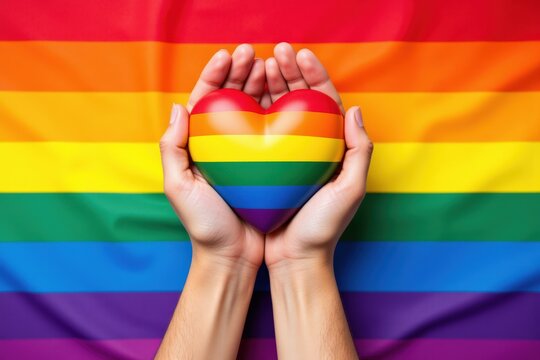 A pair of hands holding a colorful rainbow heart against a vibrant Pride flag background, symbolizing love, diversity, and LGBTQ+ acceptance.