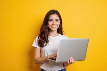 Naklejka premium Smiling Young Woman in Casual Clothing Using Laptop Against Bright Yellow Background, Symbolizing Digital Communication and Modern Workspaces