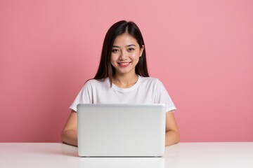 Naklejka premium Young Asian Woman in Casual White T-Shirt Working on Laptop at Home with Pink Background, Smiling Cheerfully and Engaged in Her Online Activities
