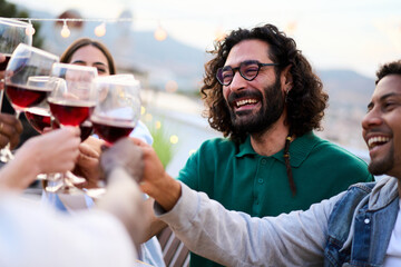 Meeting of millennial people smiling toasting with red wine glasses celebrating rooftop summer party. Young joyful friends together holding alcoholic beverages enjoying free time on weekend outdoors
