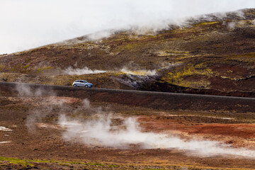 Scenic Drive Through Breathtaking Geothermal Landscape with Steam and Unique Terrain, Showcasing Natural Wonders and a Vehicle's Journey on a Serene Road