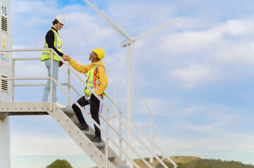Two multiracial engineers shaking hands on metal stairs of wind turbine, celebrating successful renewable energy project