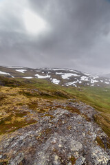 A Dramatic Landscape of Rugged Mountains Under Overcast Skies with Snow-Capped Peaks and Vibrant Green Hills in the Foreground Captured from a Unique Perspective