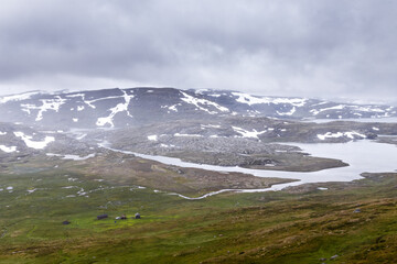 A Stunning View of Norwegian Highlands Featuring Serene Waters, Snow-Capped Mountains, and Verdant Meadows Under Dramatic Cloudy Skies Captured in a Recently Taken Image