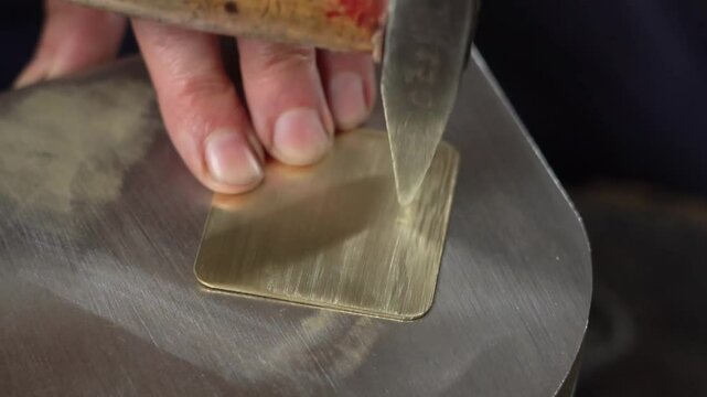 Close-up of a hand engraving a brass plate using a metal stamping tool on a steel surface. The process creates a textured pattern on the metal.