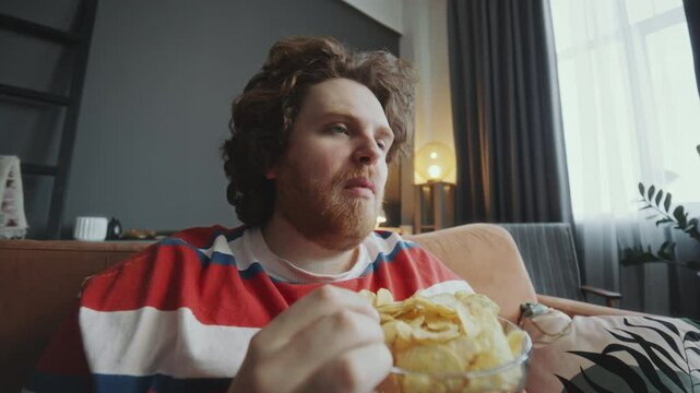 Young obese man relaxing on couch, eating potato chips from bowl and watching TV during day at home. Whip zoom shot