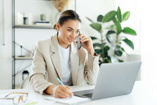 Positive busy businesswoman, office employee in a beige suit is talking on the phone while writing notes at her desk, showing professionalism and effective communication in her work environment