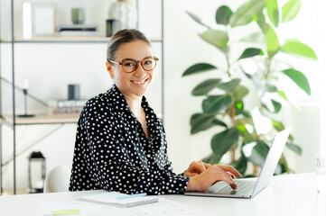 Happy pretty caucasian businesswoman wearing glasses, typing on a laptop at her desk in a bright...