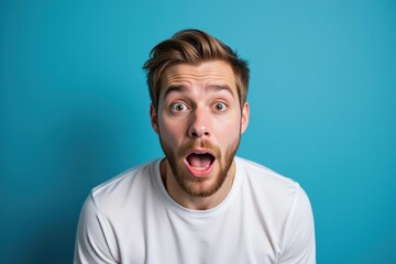 A surprised young Caucasian man with a beard, dressed in a white t-shirt against a vibrant blue background, expressing shock and excitement with his mouth wide open and eyes wide.
