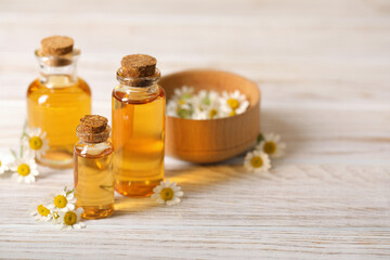 Bottles of essential oil and chamomile flowers on white wooden table, closeup. Space for text