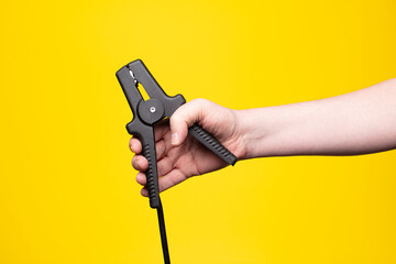 Hand holding black jumper clamp on yellow background – car battery concept, close-up.
