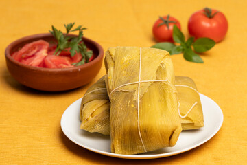 un par de humitas envueltas en hojas de choclo en un plato blanco  junto a un plato de greda con ensalada de tomate decorada con hojas verdes, atrás tomates rojos y hojas verdes, fondo amarillo