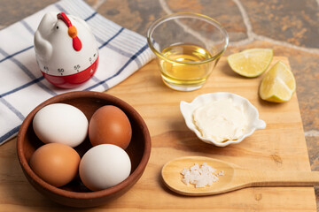 un plato de greda con huevos blancos y de color café, una cuchara con sal, un plato con mayonesa, un vaso con aceite, unos trozos de limón, un reloj de cocina y un paño de cocina