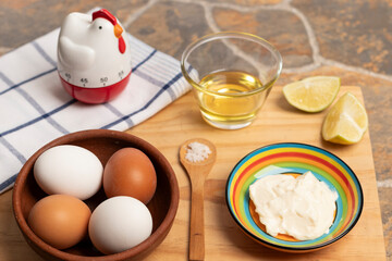 un plato de greda con huevos blancos y de color café, una cuchara con sal, un colorido plato con mayonesa, un vaso con aceite, unos trozos de limón, un reloj de cocina y un paño de cocina