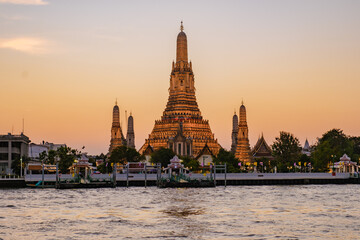 Naklejka premium As the sun sets over Bangkok, Wat Arun stands majestically by the river, bathed in golden light. Visitors admire the stunning architecture and vibrant colors, creating a serene atmosphere at dusk.