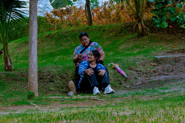 Fototapeta premium A mother and her teenage girl are sitting casually under a tree in a serene garden park, enjoying a cheerful conversation, surrounded by lush greenery.