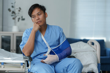Pensive doctor wearing arm sling sitting on wheelchair in hospital room