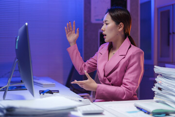 Young asian businesswoman in a pink suit experiencing stress during a late-night video call in the...