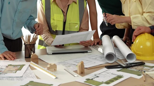 Smart architect and safety engineer discussion about building blueprint on table with architectural document, measurement tools and wooden block scatter around at modern office. Closeup. Delineation.