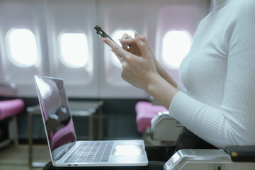 Fototapeta premium A businesswoman is sitting at the airplane window typing on her laptop while traveling. She is focused and relaxed. She is able to balance work and leisure, which is a symbol of independence.