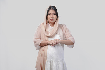 Upset Asian Muslim woman wearing headscarf veil checking time on hand gesture.