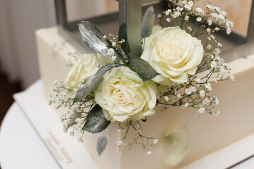 A bouquet of white roses, decorated with small white gypsophila flowers and green leaves with a silver coating. A corner of the white cake is visible in the background.