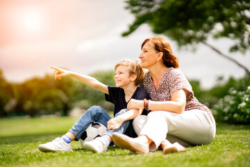 Fototapeta premium Healthy and happy grandmother sitting on green grass with joyful grandchild at summer park look away. Joyful cuacacian kid take a rest affter exercise with senior woman at garden.