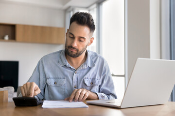 Man engaged in financial or administrative work seated at table at home office, using calculator, pay bills through e-banking system on laptop, review documents, busy in personal finances management