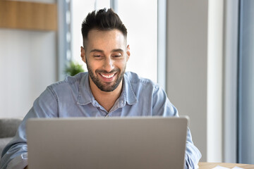 Smiling Latin man using laptop at home office, working remotely, studying or learning new educational platform, enjoy pleasant news received in email, buying goods and services through marketplace app