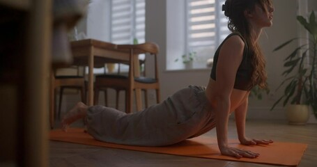 A brunette girl in a black top and gray pants does a Cobra pose while doing yoga and sports on an orange mat in the kitchen at home