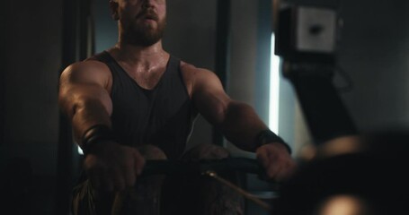 Close-up shot of a bearded man in a tank top exercising on a rowing machine in a contemporary gym