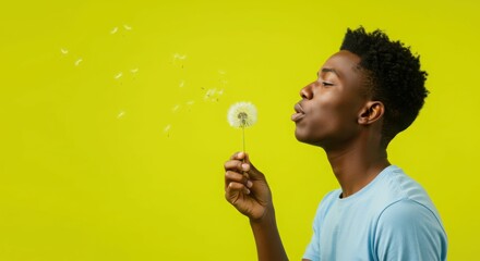 Fototapeta premium Young african male blowing dandelion on bright yellow background