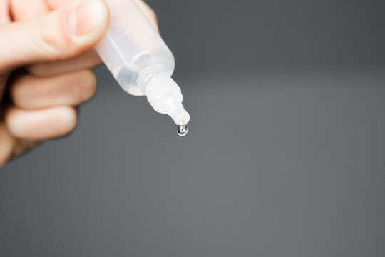 Drop hanging from the transparent tip of a small plastic eye drop or ear drop bottle just before it falls. Hand holding plastic bottle of medicine with droplet. Isolated against a gray background. 